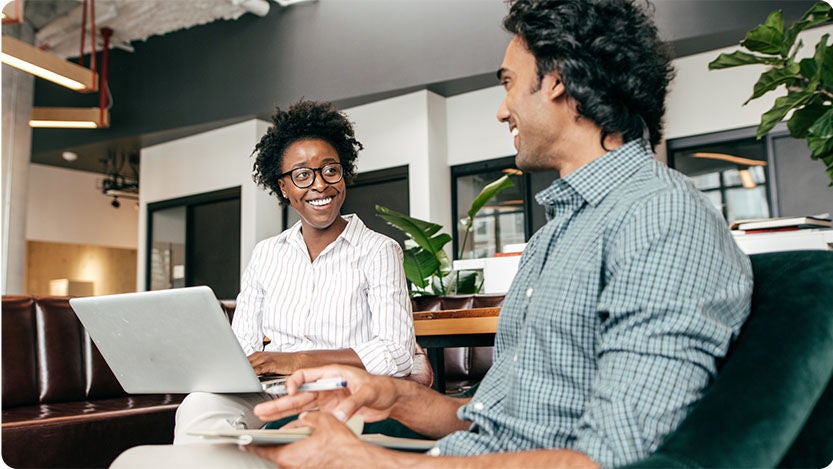 Two people sitting indoors. One person is holding a laptop and both are smiling at each other.