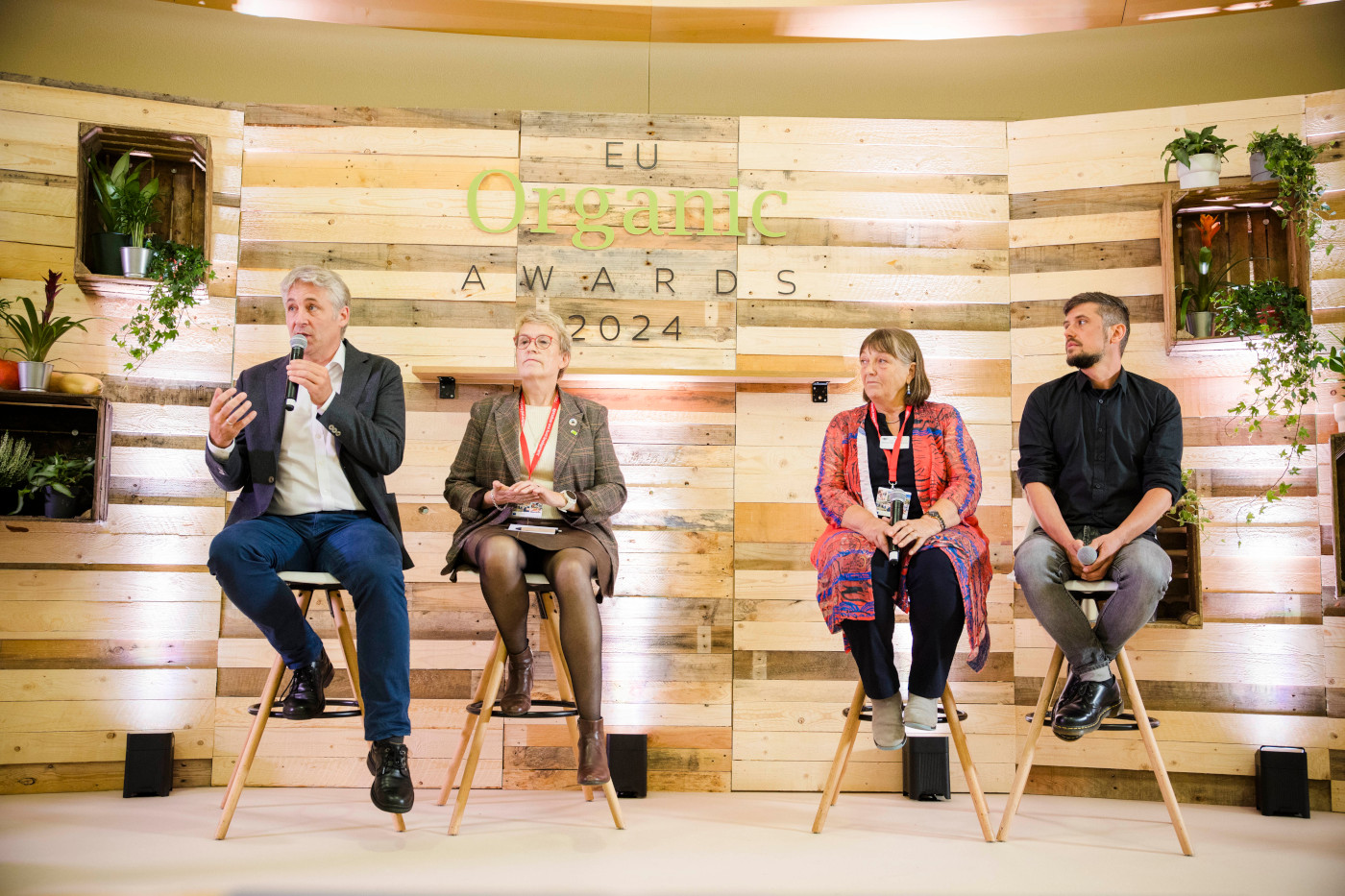 4 people sitting on chairs on a stage, a man is speaking with a microphone