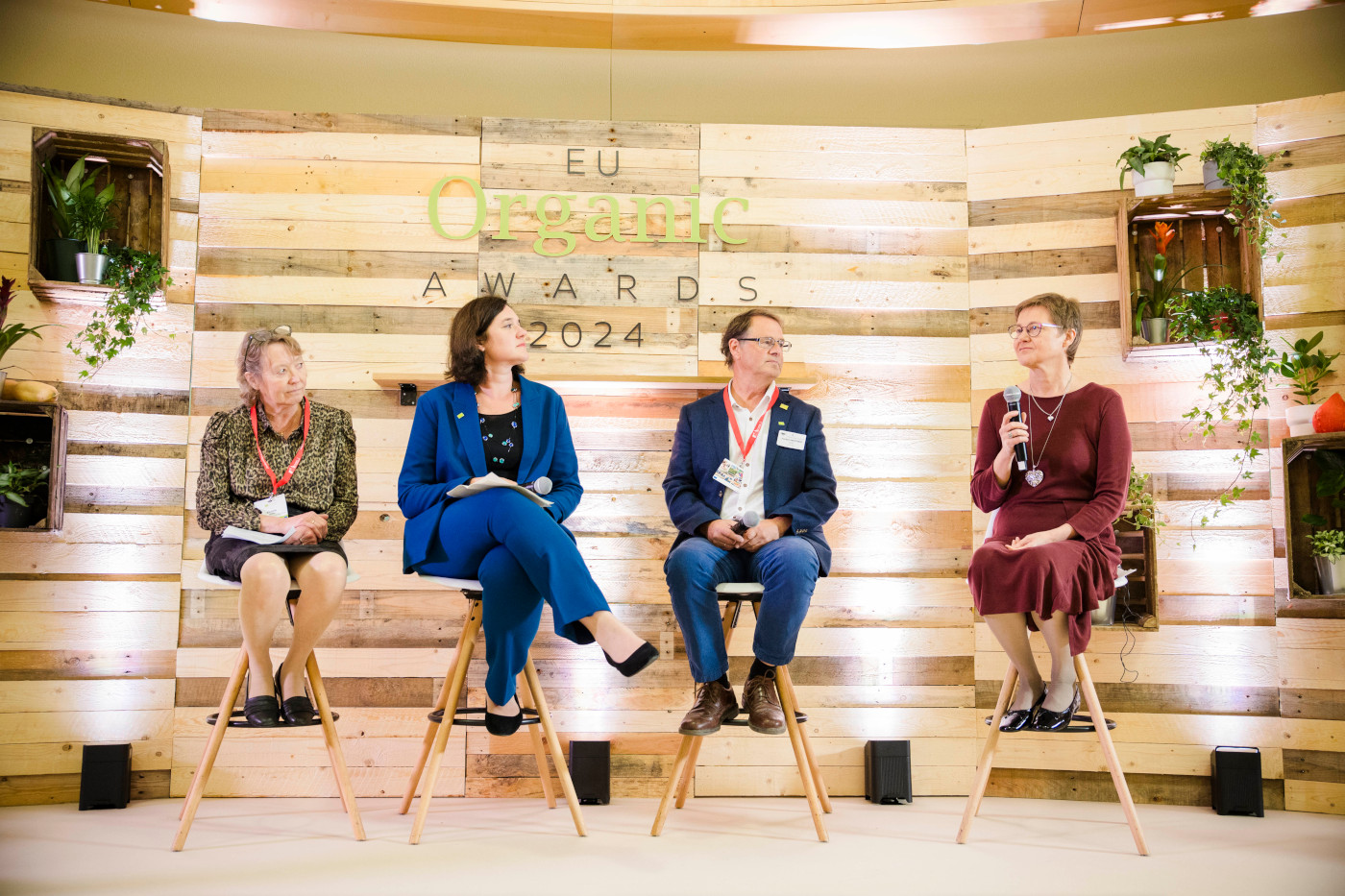 3 ladies and a man sitting on chairs on a stage, one lady holds a microphone