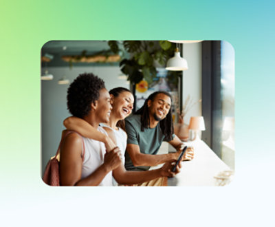 Three people smiling and looking at a tablet while sitting at a counter by the window in a bright room.