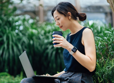 コーヒーを片手に屋外でノート PC を使う人物。