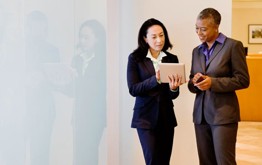 Two businesswomen dressed in suits are using a digital tablet while chatting in an office corridor. 