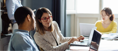 A woman with red glasses uses a laptop while talking to a man. Another woman is working on a laptop in the background.