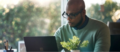 A man wearing glasses sits at a table working on a laptop, with a plant and pens nearby. 
