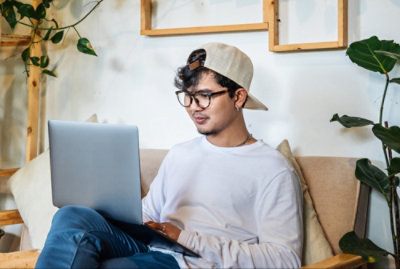 Person wearing glasses and a cap sits on a couch using a laptop, surrounded by plants.