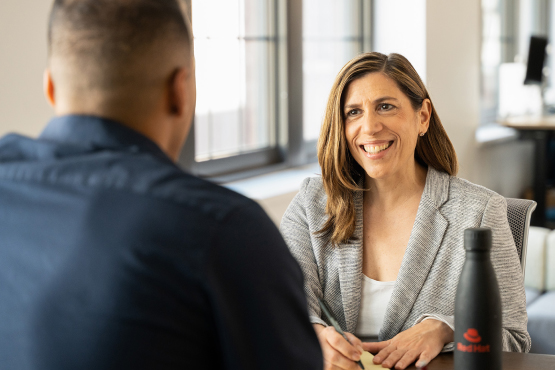 Close up of two people sitting at a table talking. The angle of the image is from behind the head of the person in the foreground. Both people are dress professionally and are enjoying the conversation. 