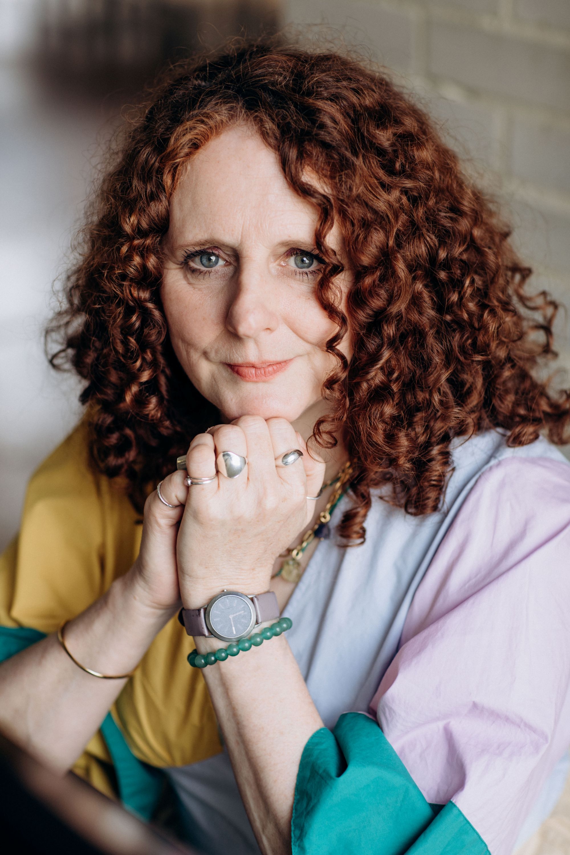 A woman with red curly hair resting her chin on her hands, wearing rings on her fingers and a watch.