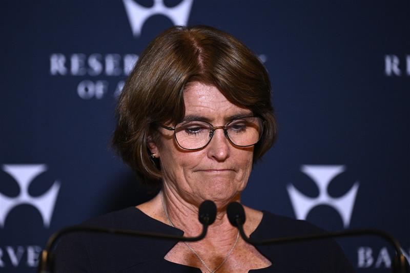 A dark-haired woman in glasses casts her gaze downward as she stands at a lectern in forrnt of an RBA backdrop.