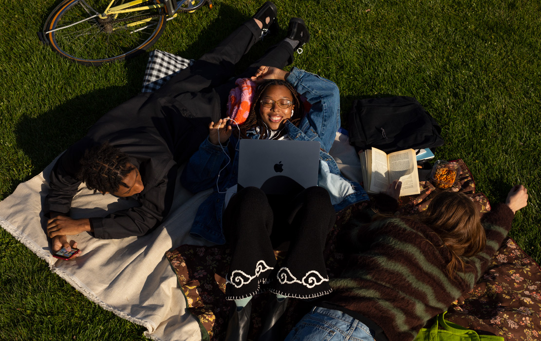 Three uni students lie on a blanket in the park. One student has an iPhone. One student has a MacBook Air and Apple headphones. One student has a paperback book.