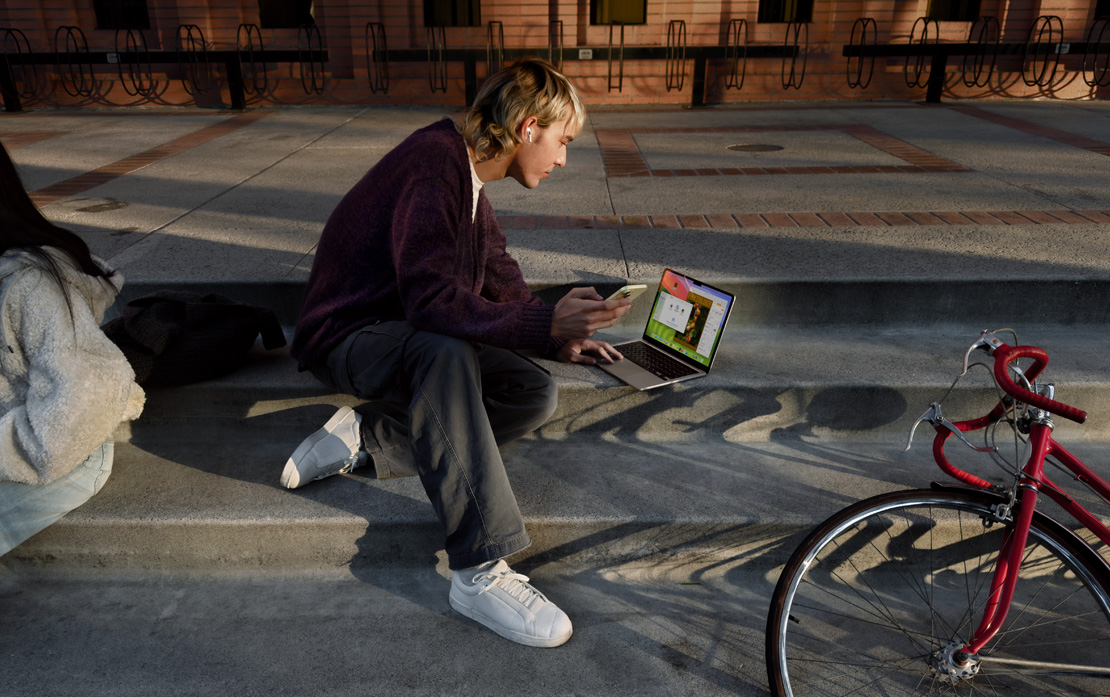 A uni student sitting on steps with a bicycle using an iPhone and MacBook