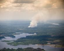 Aerial view of smoke rising from a large fire in a forested area near a river.