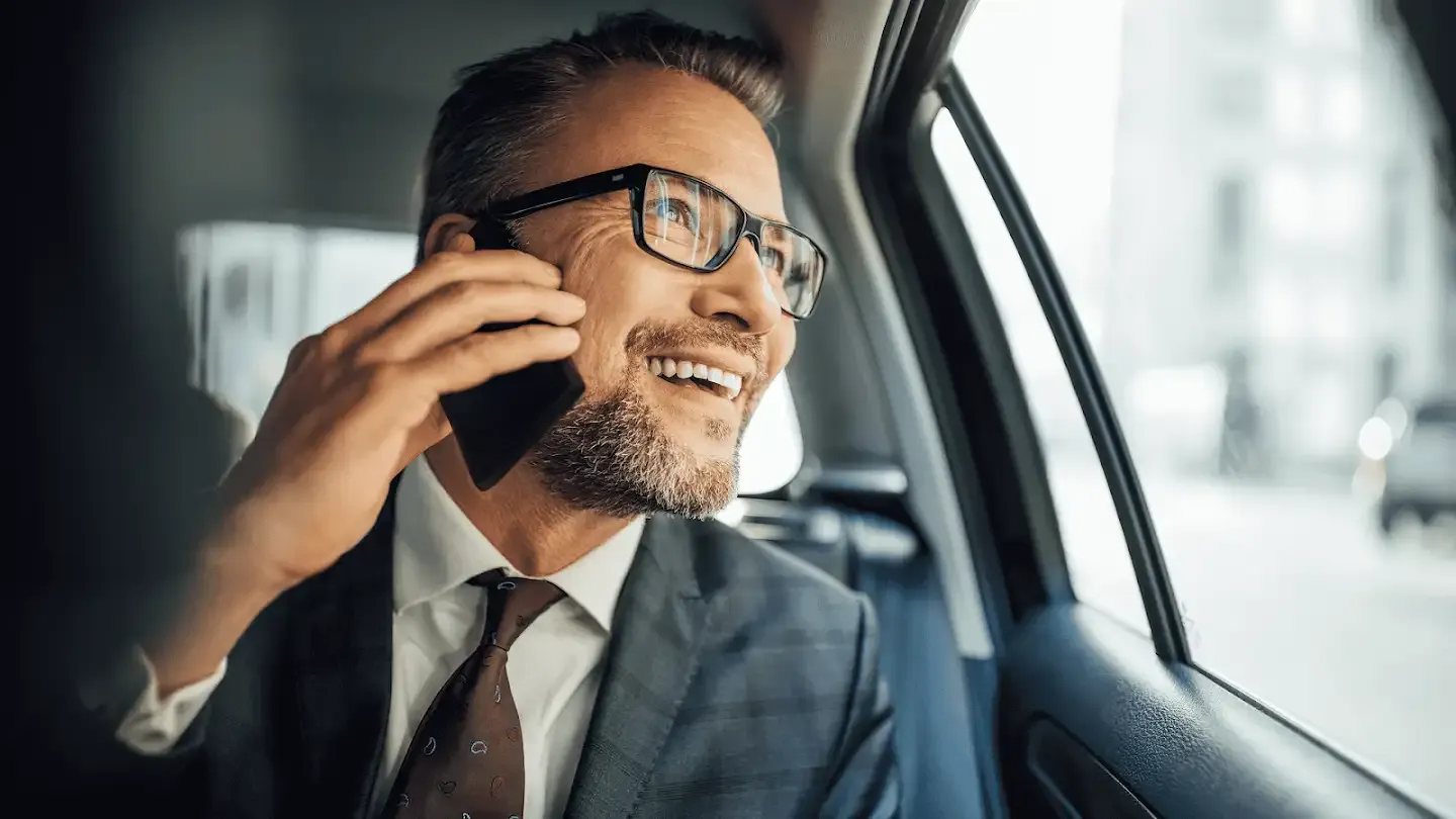 Un hombre blanco con un traje de cuadros grises y corbata color granate habla por su teléfono móvil mientras mira por la ventana en el asiento trasero de un coche.