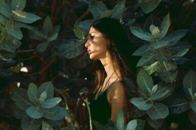 A woman stands in profile with her face partially obscured by shadows, surrounded by thick green leaves, representing the hidden nature of cutting and self-harm.