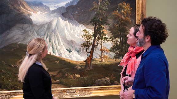 Two women and a man in front of a large painting of a glacier. 