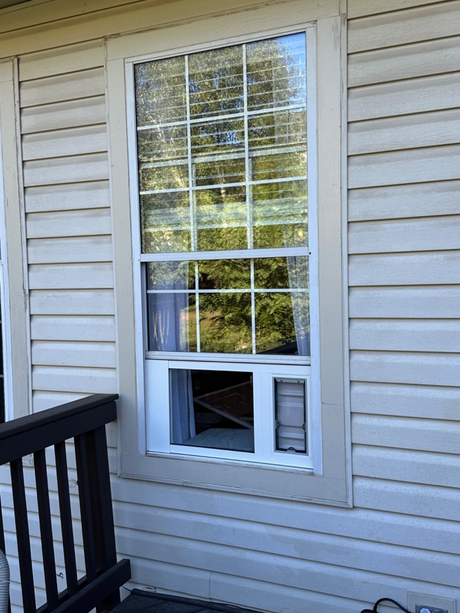 Liberty Doggie Door for sash window mounted in the lower section of a white sliding glass door, the upper section showing a 2x3 grid of glass panes with outdoor landscape visible.