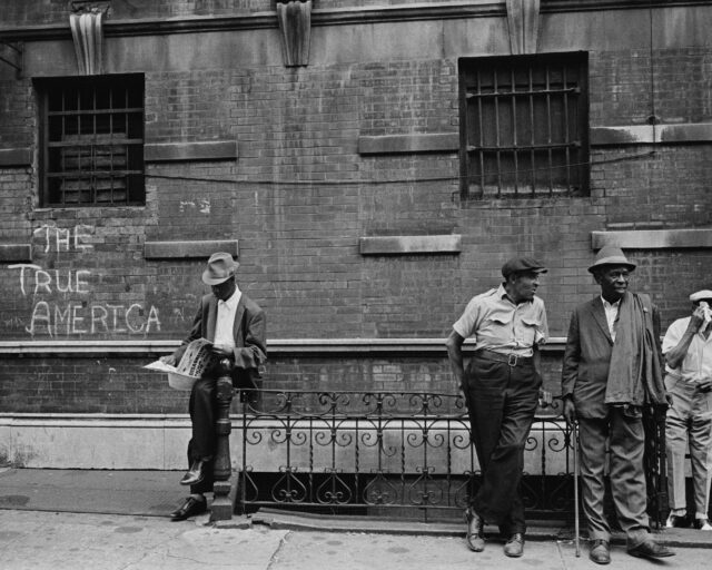 Four men in 60s-era clothing stand on a sidewalk. Written on the building behind them is