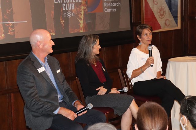 Three people sit on stools in front of an audience with a presentation screen behind them.