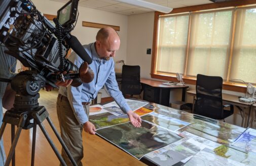 Greg Fiske displays printed out maps on a table to a camera crew