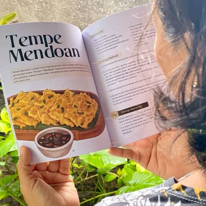 Une femme, tenant dans ses mains le livre de cuisine « Tempe and Nasi Campur », ouvert sur une recette de Tempe Mendoan avec des illustrations dessinées à la main.