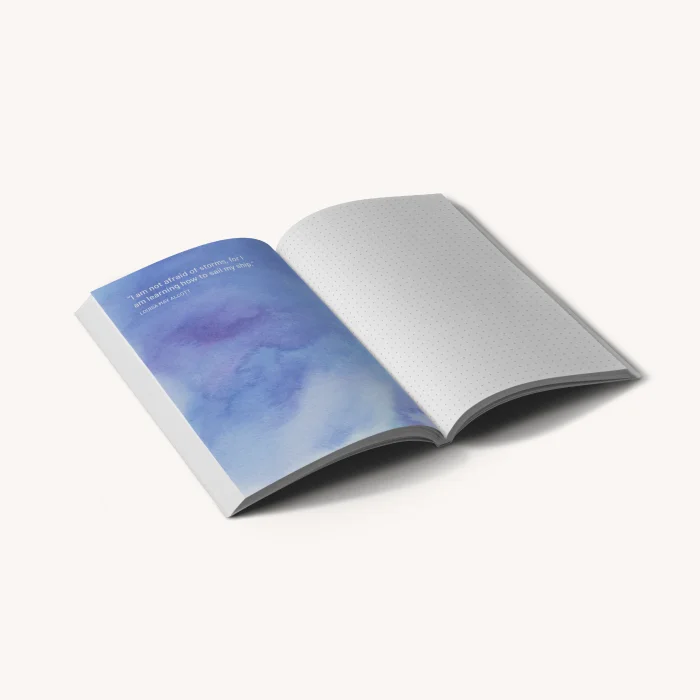 Cuaderno de tapa blanda abierto con una página pintada en acuarela azul y otra con cuadrícula de puntos.