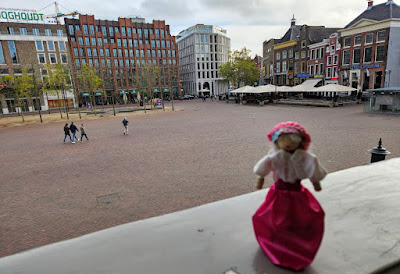 Etta Palm op bordes het poppetje van Etta Palm op de rand van het bordes van het Groninger Stadshuis met op de achtergrond nieuwe gebouwen aan de oostkant van de Grote Markt en rechts oudere gebouwen