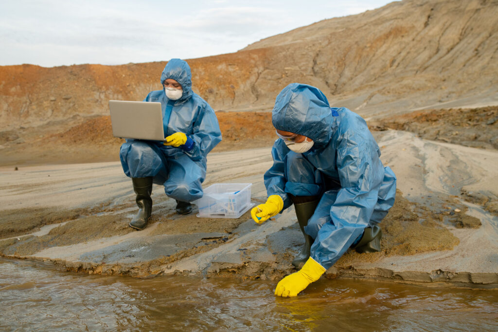Two scientist wearing protective suit in a barren landscape taking water sample from a small stream.