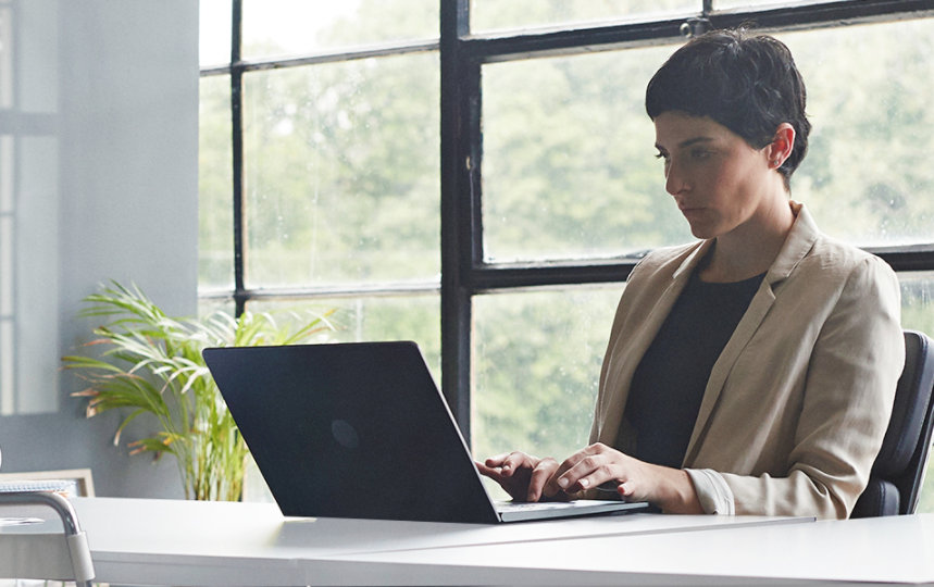 A woman is sitting at a long desk and typing on a laptop in a modern window office that has glass walls.