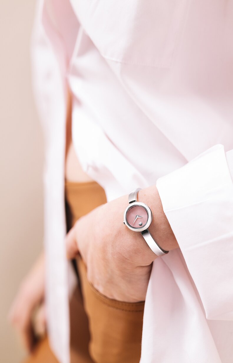 Woman in a white shirt and brown pants with her hand in her pocket wearing a silver Museum Bangle watch.