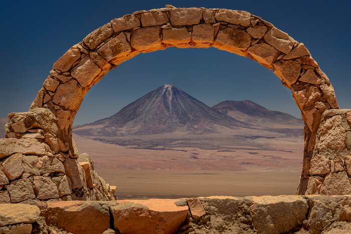 An old-looking arch made of imperfectly-shaped terracotta stones stands in the foreground, framing the view on a sunny desert landscape with a volcano. The bright blue sky contrasts with the ground’s red, orange and brown colours. The Licancabur volcano, in the centre, stands next to a smaller mountain, both have darker colours, leaning towards purple and green. The top of the volcano has hints of white.