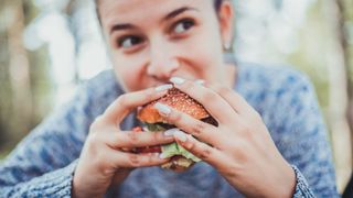 Woman is shown smiling and eating a burger while looking into the distance