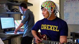 a student wearing a "drexel" shirt plays guitar while wearing an EEG cap. A researcher behind him reads brain waves from a computer screen