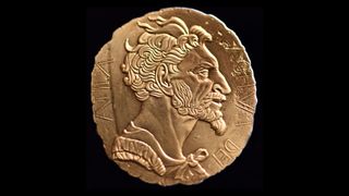 Attila the Hun's face on a gold-looking coin with his name around the edge. His face is facing right and he has lots of tuffs of short, wild hair, large ears, a strong brow, large nose, and a beard and moustache.