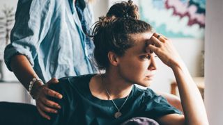 a photo of a woman sitting down with one hand on her head, as if frustrated; a person standing behind her has a hand on the woman's shoulder