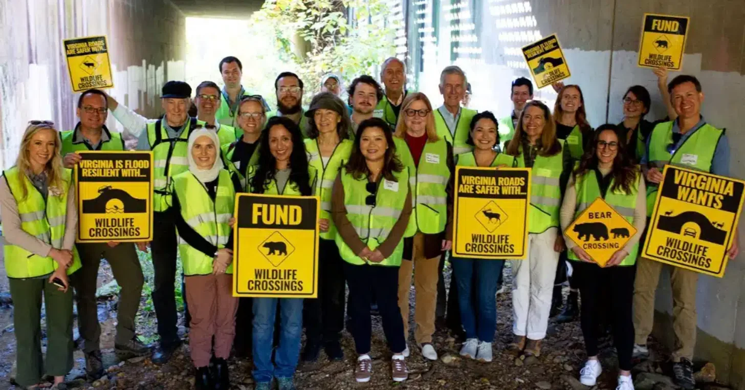 A smiling group of advocates in bright yellow safety vests holds signs promoting wildlife crossing funding, gathered indoors to support road safety initiatives for Virginia wildlife.