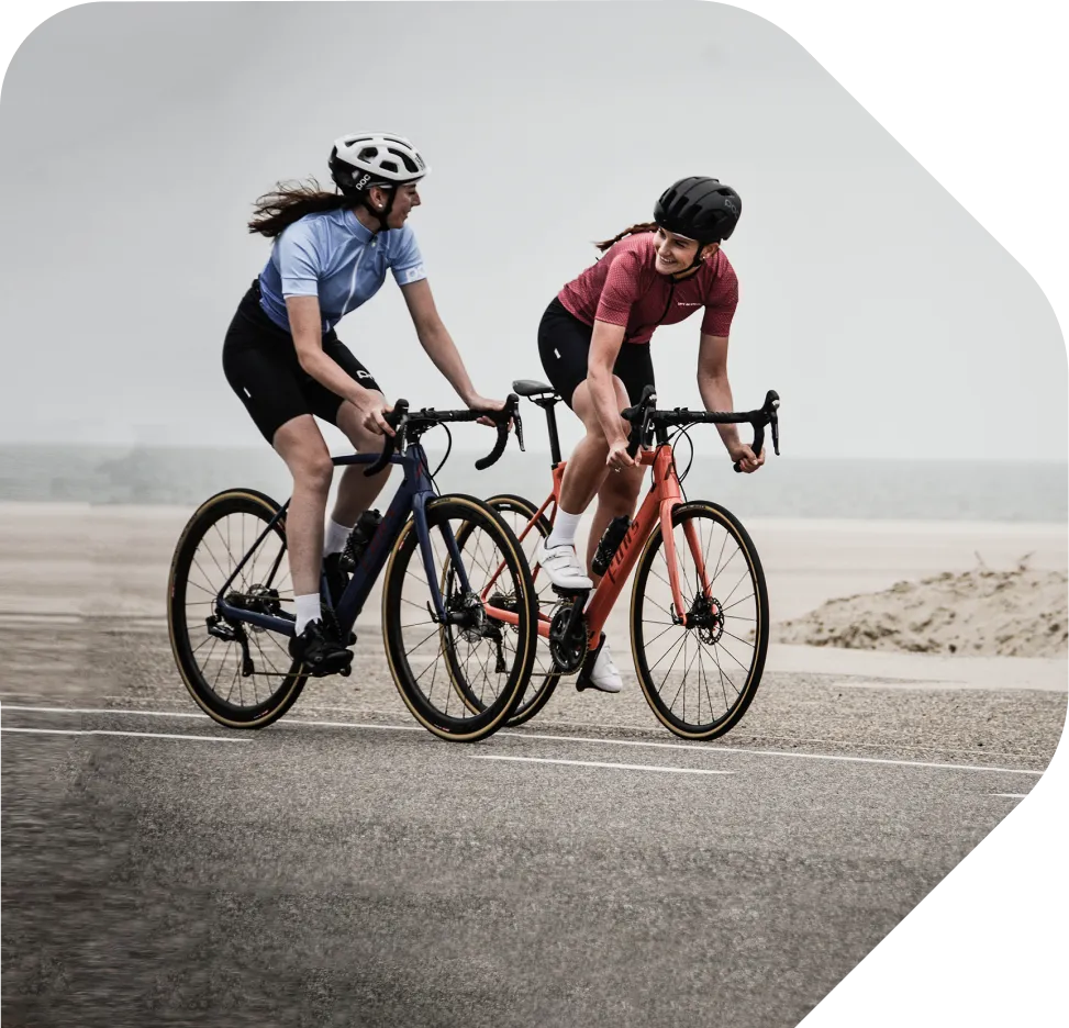 Two young women wearing lycra shorts and t-shirts cycling on a road, next to the beach. 