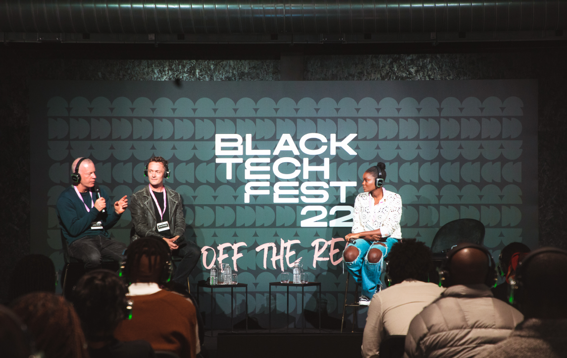 A diverse group of attendees seated at the Black Tech Fest event, with a panel of hosts engaging in discussion on stage