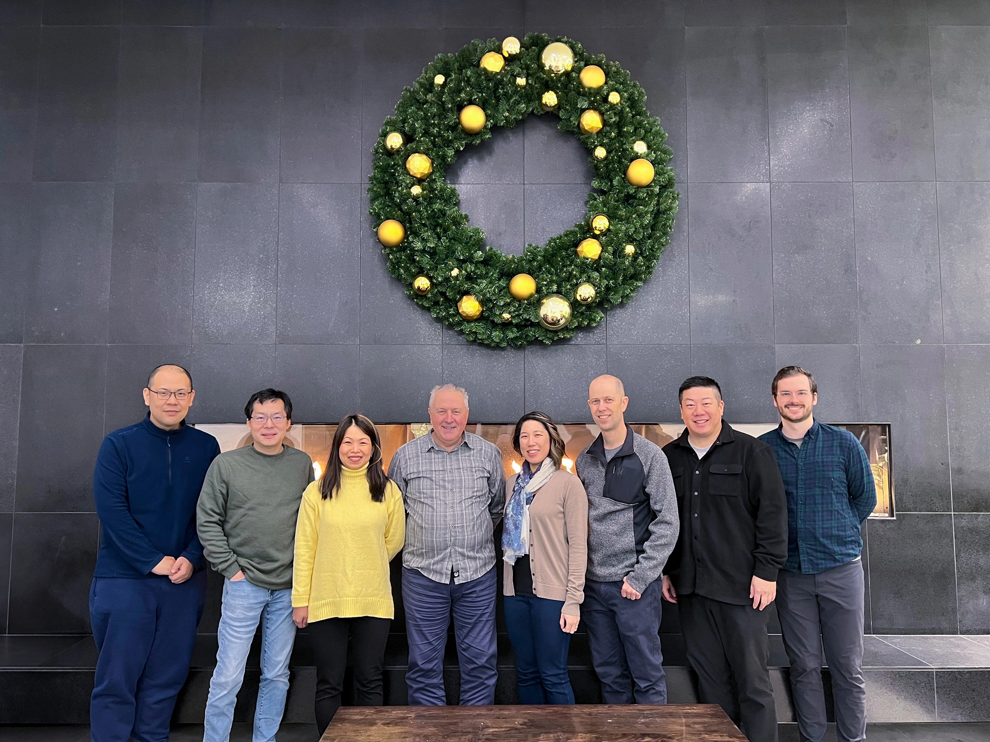 A group of 8 people standing in a line facing the camera. There's a huge wreath with yellow and gold ornaments hanging on the grey tiled wall above the group.