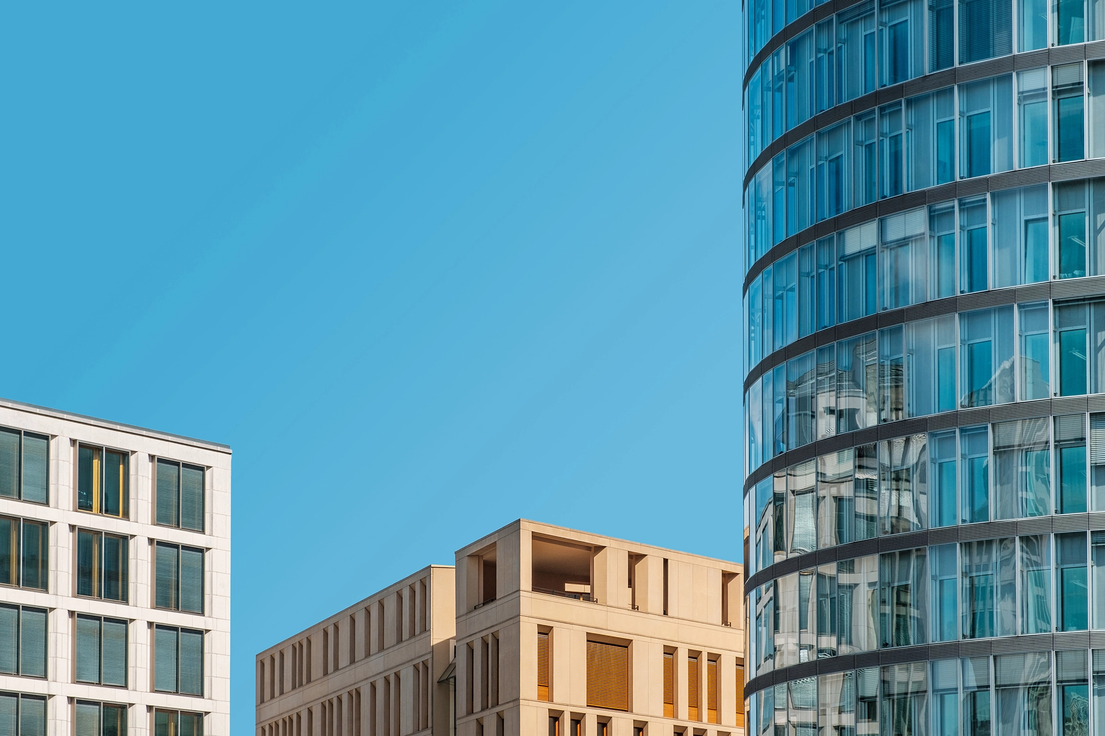 A row of three buildings against a clear blue sky. The left-most building is white-grey concrete, the middle building is sandy concrete, and the right-most building is almost all glass. The top of two are visible, and the one furthest to the right is not.