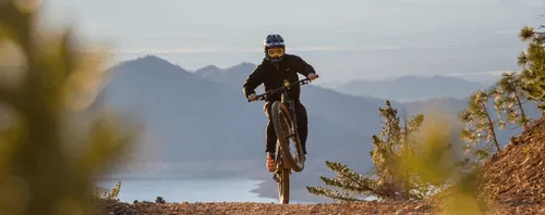 A mountain biker wearing a helmet and goggles performs a wheelie on a dirt trail with scenic mountains and a lake visible in the background.