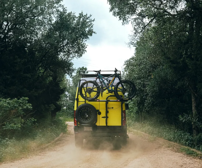 A yellow van with spot branding and two bicycles mounted on its rear drives down a dusty, tree-lined dirt road.