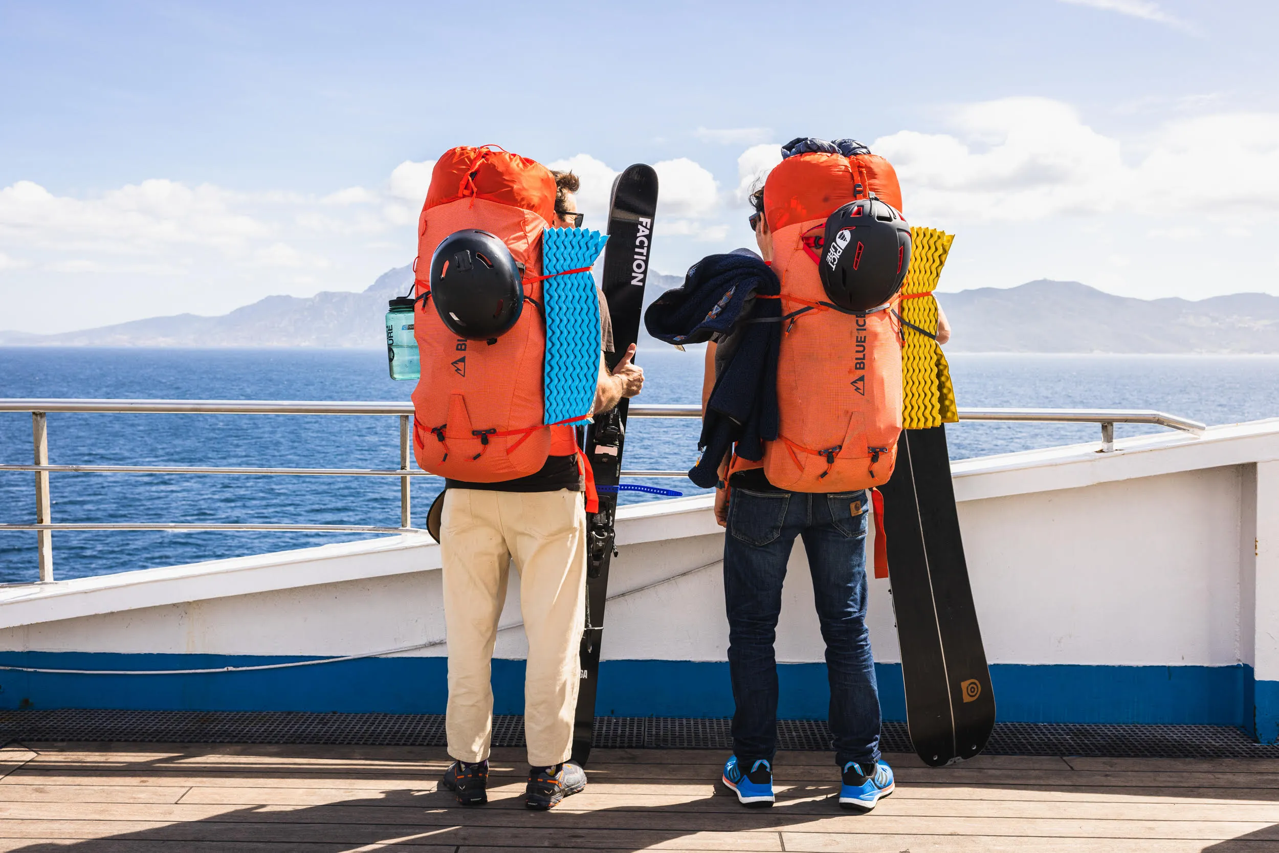 Two people with large orange backpacks and skiing equipment stand on a boat deck overlooking the ocean and distant mountains.