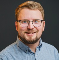 A young man wearing glasses smiles against a dark background.