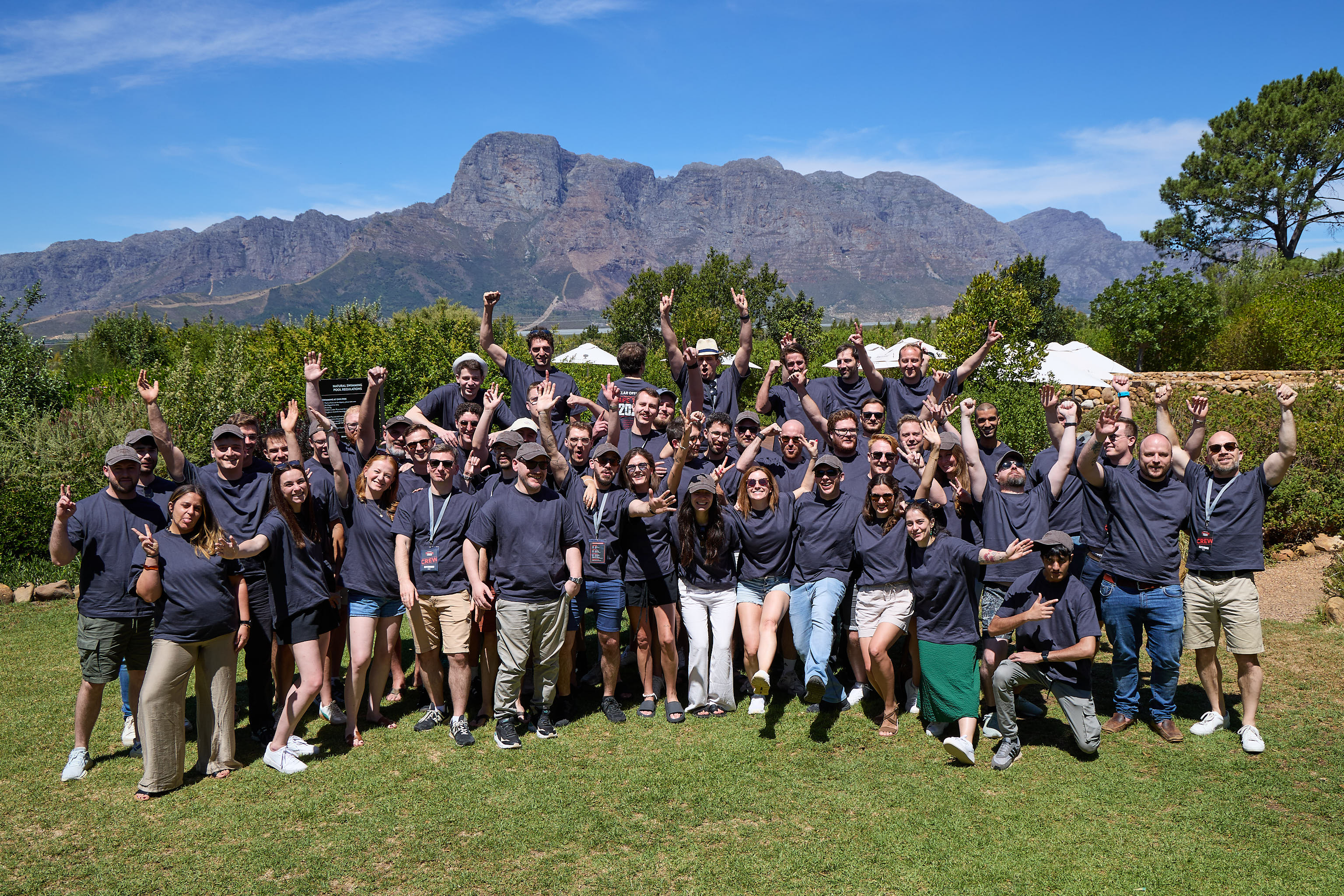 A large group of cheerful people wearing dark T-shirts in front of a mountain landscape on a sunny day.