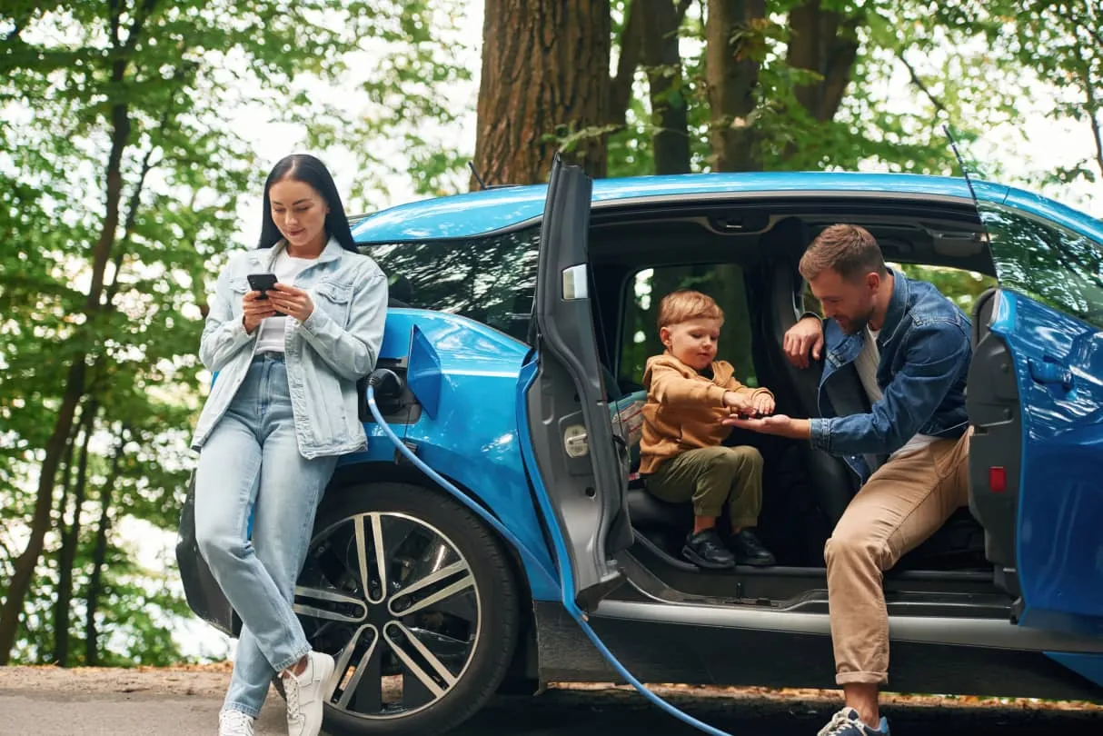 Woman standing by a blue electric car charging outdoors while a man and child sit inside the open car, interacting.