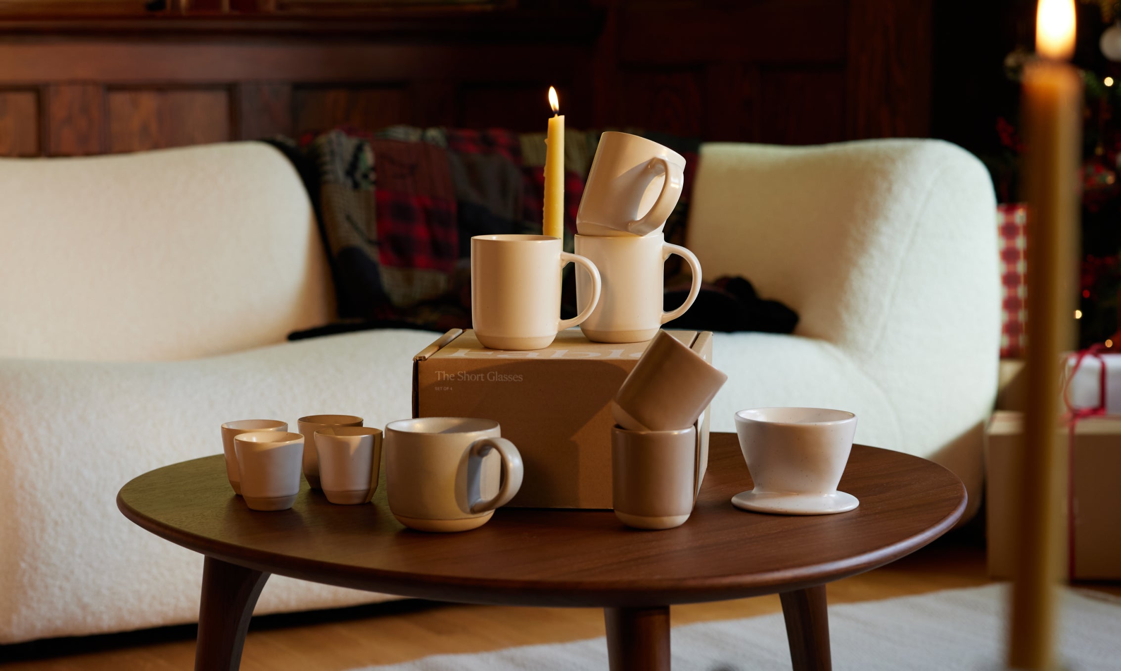 A wooden table with stacked mugs and a candle in a cozy living room setting.