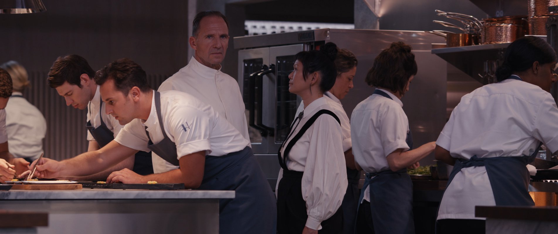 Man in a white chef’s coat stares ahead as he’s surrounded by staff, including one woman wearing an apron looking directly up at him.