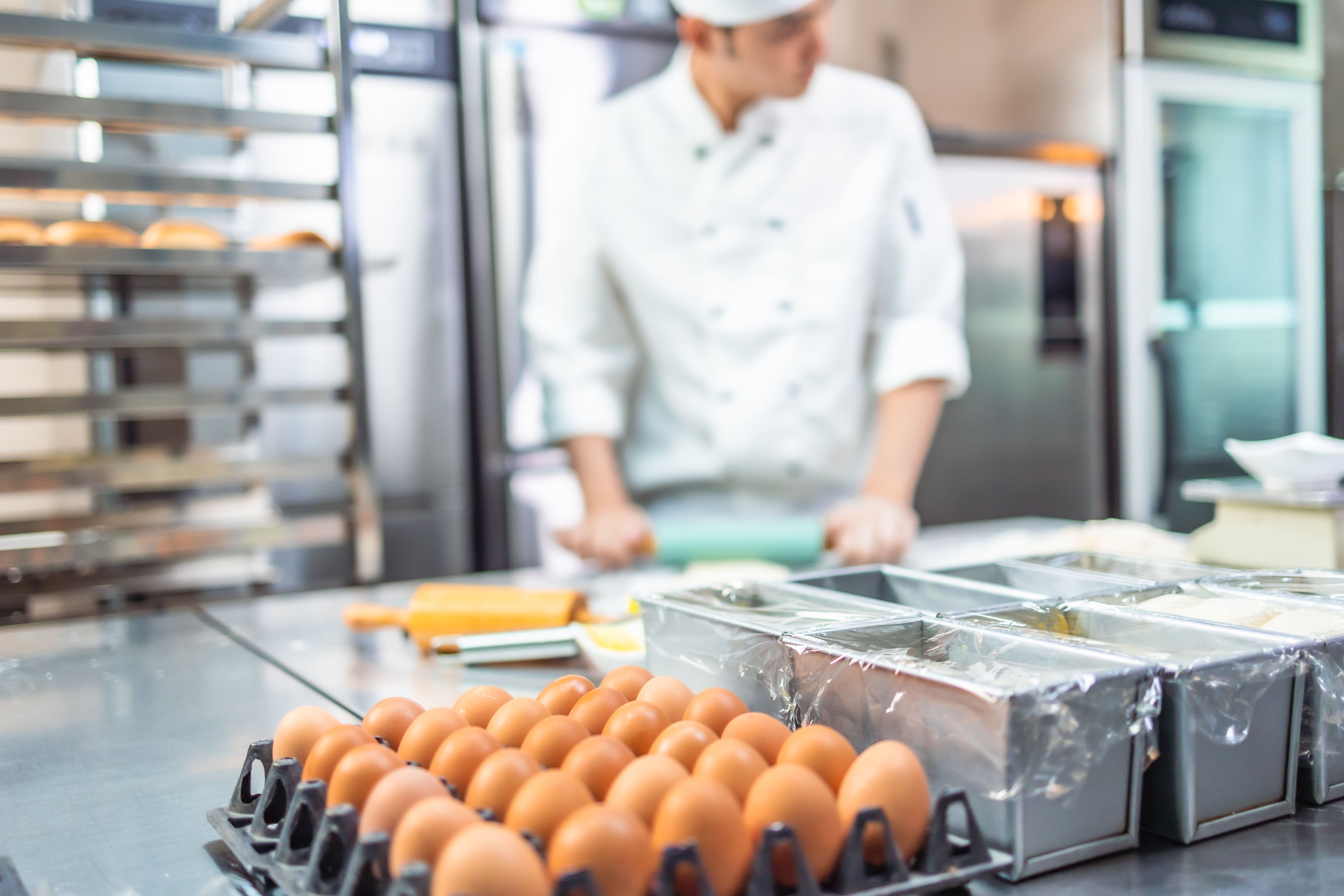 In the foreground, a carton of eggs sits on a metal countertop while a baker in whites and a chef’s hat rolls out dough in the background. 