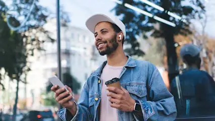 Een persoon met een telefoon en koffie staat buiten, met bomen en gebouwen op de achtergrond.