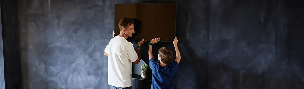 Een man en een kind plaatsen samen een bruin vierkant object aan een donkere muur.