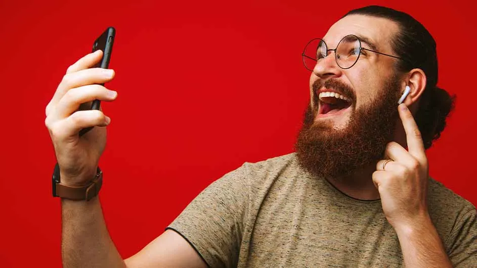 Een man met een baard, een bril en een oortje lacht en houdt een telefoon vast tegen een rode achtergrond.
